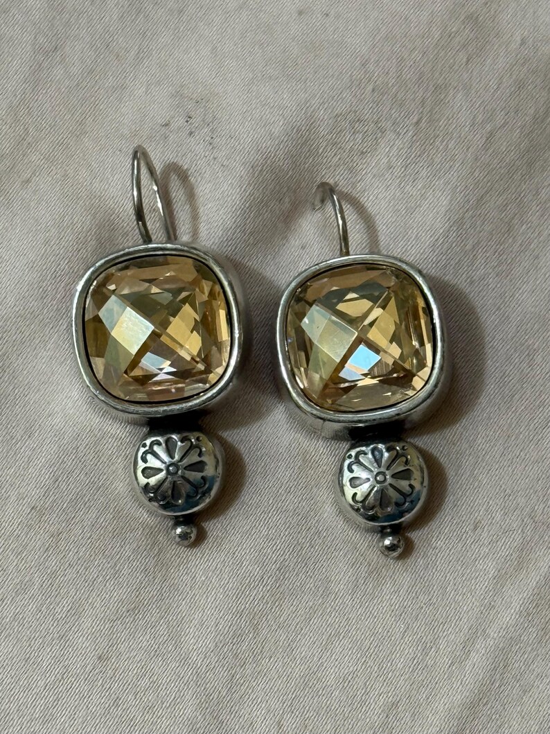May include: A pair of drop earrings featuring square-cut, light amber-colored faceted crystals set in silver-toned metal. Below each crystal is a small, round silver-toned bead with an embossed floral pattern and a tiny spherical tip at the base.