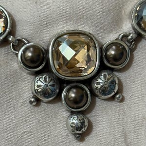 May include: A necklace featuring a central square-cut champagne-colored crystal surrounded by dark metallic beads and silver-toned circular charms with etched floral patterns. The design includes additional faceted crystals on the sides.