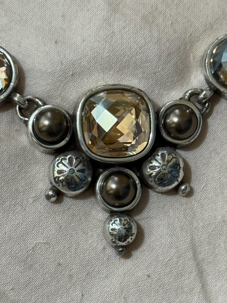 May include: A necklace featuring a central square-cut champagne-colored crystal surrounded by dark metallic beads and silver-toned circular charms with etched floral patterns. The design includes additional faceted crystals on the sides.