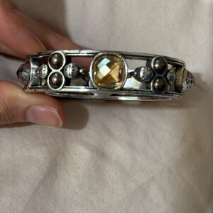 May include: A silver-toned metal bangle bracelet featuring a central faceted amber-colored stone, flanked by clusters of small dark brown spherical beads and clear faceted crystals set within a structured, open-work frame design.