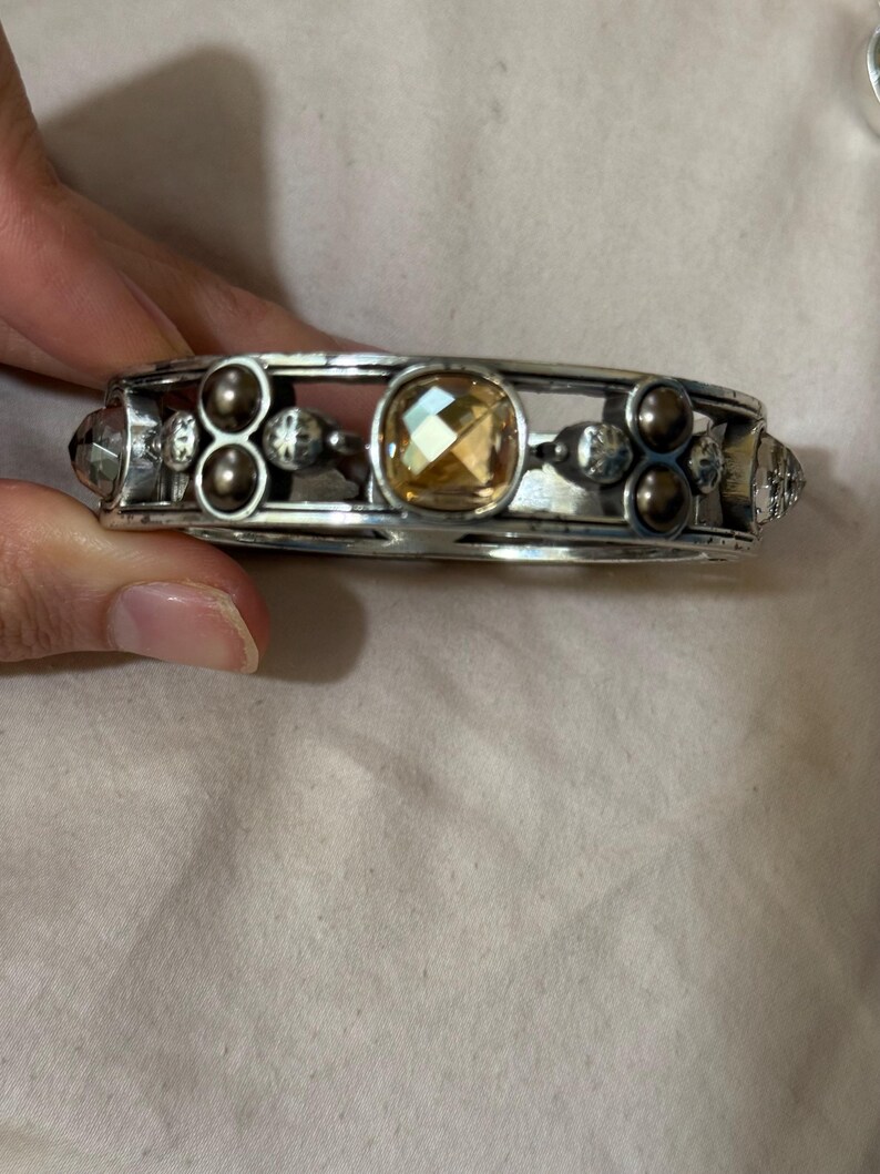 May include: A silver-toned metal bangle bracelet featuring a central faceted amber-colored stone, flanked by clusters of small dark brown spherical beads and clear faceted crystals set within a structured, open-work frame design.