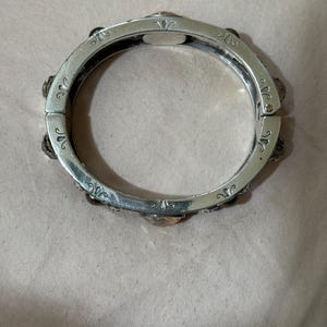 May include: A silver-toned hinged bangle bracelet featuring a series of evenly spaced, faceted amber-colored stones set into the metal band. The surface of the bracelet is decorated with engraved scrollwork patterns between each stone setting.