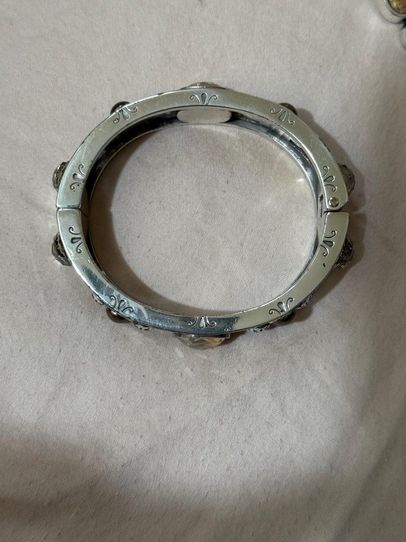 May include: A silver-toned hinged bangle bracelet featuring a series of evenly spaced, faceted amber-colored stones set into the metal band. The surface of the bracelet is decorated with engraved scrollwork patterns between each stone setting.