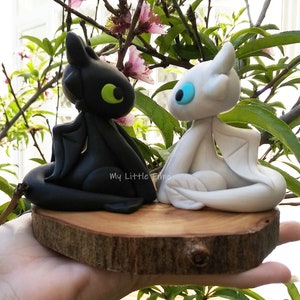 May include: Two small dragon figurines, one black with green eyes and one white with blue eyes, are sitting on a wooden base. The base is on a person's hand. The text "My Little Fimo" is visible on the base.