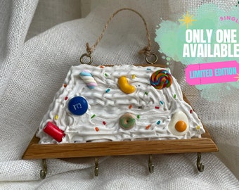 Puede incluir: Un porta llaves de madera blanco, crema y marrón con forma de porción de pastel con varios adornos de caramelo, incluyendo un bastón de caramelo, un caramelo amarillo, una piruleta, una dona y un M&M. El porta llaves tiene un asa de cuerda y tres ganchos.