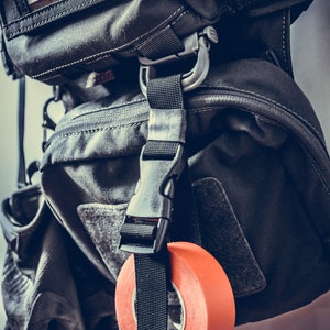 Könnte beinhalten: Ein schwarzer taktischer Rucksack mit einem schwarzen Gurt und einer Rolle orangefarbenem Klebeband. Der Rucksack hat ein Etikett mit der Aufschrift "STAFF #82".