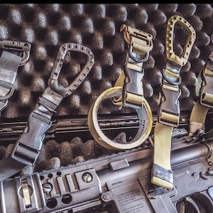 May include: Five different types of tactical slings with black and tan webbing and metal clips. The slings are arranged on a black foam surface with a roll of tan tape and a black rifle.