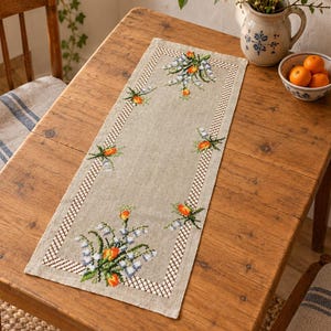 May include: A rectangular beige linen table runner featuring a cross-stitched border pattern and floral motifs in shades of orange, green, and white. The embroidery depicts clusters of lily of the valley flowers with vibrant orange accents.