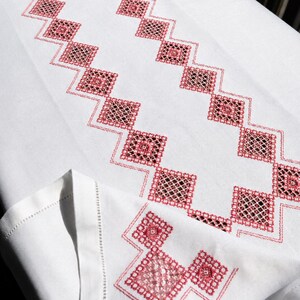 Puede incluir: Un camino de mesa rectangular blanco con un patrón geométrico central de diamantes interconectados bordados en hilo rojo. El diseño incluye intrincados detalles de punto de cruz dentro de cada forma de diamante, con un motivo a juego en la esquina.