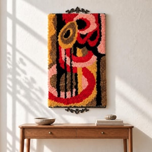 May include: A rectangular wall hanging featuring a high-pile tufted texture with an abstract pattern of concentric circles and vertical lines in shades of red, pink, mustard yellow, brown, and black, mounted on a dark metal frame with ornate handles.