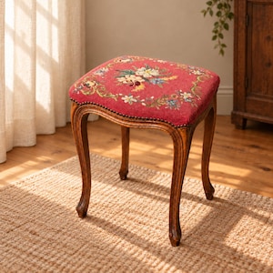 Puede incluir: Un taburete cuadrado con un asiento de tela roja que presenta un patrón floral de tapiz multicolor. El taburete tiene cuatro patas de madera curvadas con un acabado marrón oscuro y un borde decorativo de tachuelas metálicas en el asiento.