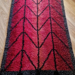 May include: A rectangular shag rug with a geometric design. The rug features a gradient of red hues, with black geometric lines and a black border. The rug has a fringe on the short ends.