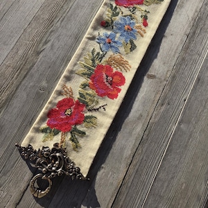 May include: Cream-colored embroidered runner featuring vibrant red poppies and blue flowers. The floral design is accented with green leaves and stems. The runner has ornate metalwork at each end, adding a touch of elegance to the handcrafted textile.
