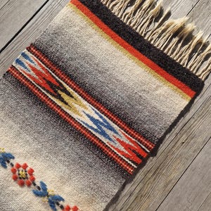 May include: A rectangular woven rug with a cream, gray, and black gradient design. The rug features a red, yellow, and black stripe, and a decorative border with red, blue, and yellow geometric patterns. The rug has cream-colored fringe.