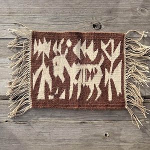 以下が含まれることがあります： 茶色の背景とオフホワイトの幾何学模様が特徴の、手織りの長方形の織物。 デザインには、様式化された動物の姿と抽象的な形が含まれています。 エッジには長いベージュのフリンジが付いています。