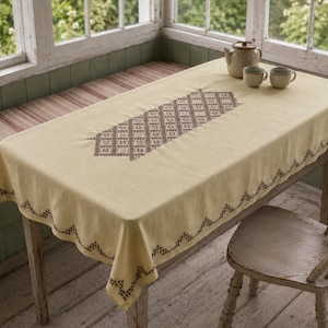 Puede incluir: Un mantel rectangular de color amarillo pálido con un patrón central de punto de cruz geométrico marrón y un borde en zigzag a juego en los extremos. La tela cubre una mesa de madera, con una tetera de cerámica y dos tazas pequeñas encima.