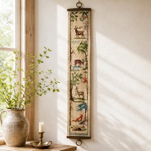 May include: A long, vertical wall hanging tapestry featuring a series of embroidered woodland scenes. The design includes a stag, a deer, and a fox amidst trees and foliage, all rendered in natural earth tones on a cream-colored fabric background.