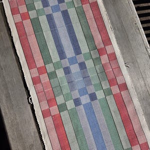 May include: A rectangular woven table runner with a colorful striped pattern. The runner features alternating bands of red, green, and blue, with a central blue checkered design. The edges are finished with a white border.