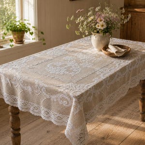 May include: A rectangular beige tablecloth featuring intricate white lace borders and a central lace panel. The fabric has a textured weave, and the lace displays detailed floral and geometric patterns along the edges and the middle section.
