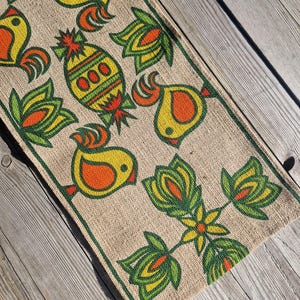 Op de afbeelding: Een rechthoekige tafelloper van jute met een herhalend patroon van gestileerde gele vogels, oranje accenten en groene en gele bloemmotieven. De loper heeft een groene rand.