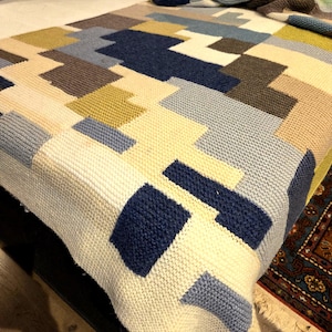 May include: A handmade, knitted blanket with a geometric pattern in shades of cream, navy blue, light blue, olive green, and brown. The blanket is draped over a bed, showcasing its textured surface and color block design. The blanket appears soft and cozy.