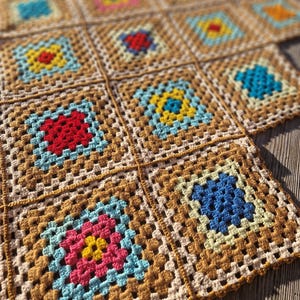 Puede incluir: Una manta de ganchillo hecha a mano con un diseño de patchwork. Cada cuadrado es de un color diferente, incluyendo rojo, azul, amarillo y rosa, sobre un fondo marrón claro. La manta está hecha de hilo y tiene un aspecto texturizado.