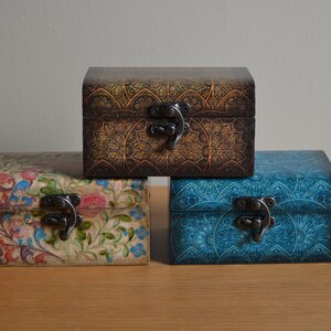 Puede incluir: Tres cajas de madera decorativas con diseños intrincados y cierres de metal. La caja superior tiene un diseño marrón y dorado, la caja del medio tiene un diseño floral y la caja inferior tiene un diseño azul y blanco.