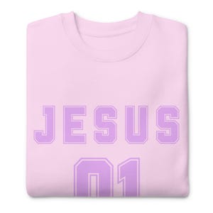 Könnte beinhalten: Hellrosa Sweatshirt mit dem Wort "JESUS" in großen, lavendelfarbenen Blockbuchstaben auf der Brust. Darunter die Zahl "01" in Lavendel. Das Sweatshirt ist ordentlich gefaltet.