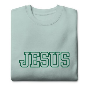 Könnte beinhalten: Hellblaues Sweatshirt mit dem Wort "JESUS" in grünen Blockbuchstaben auf der Vorderseite. Das Sweatshirt ist sauber gefaltet und zeigt das Design. Die Schriftart hat einen College-Stil.