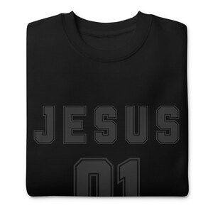 Könnte beinhalten: Schwarzes Sweatshirt mit dem Wort "JESUS" in großen, grauen Blockbuchstaben auf der Brust. Darunter steht die Zahl "01" in denselben grauen Blockbuchstaben. Das Sweatshirt ist aus weichem Material.