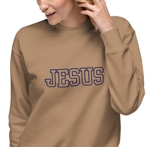 Könnte beinhalten: Ein beigefarbenes Sweatshirt mit dem Wort "JESUS" in marineblauen Blockbuchstaben. Das Sweatshirt hat einen Rundhalsausschnitt und der Text ist hellblau umrandet.