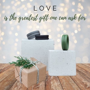 May include: A brown gift box with twine and green sprigs on top, sitting on a wooden surface. Two small concrete blocks with silicone rings on top. The text "LOVE is the greatest gift one can ask for" is written above the blocks.