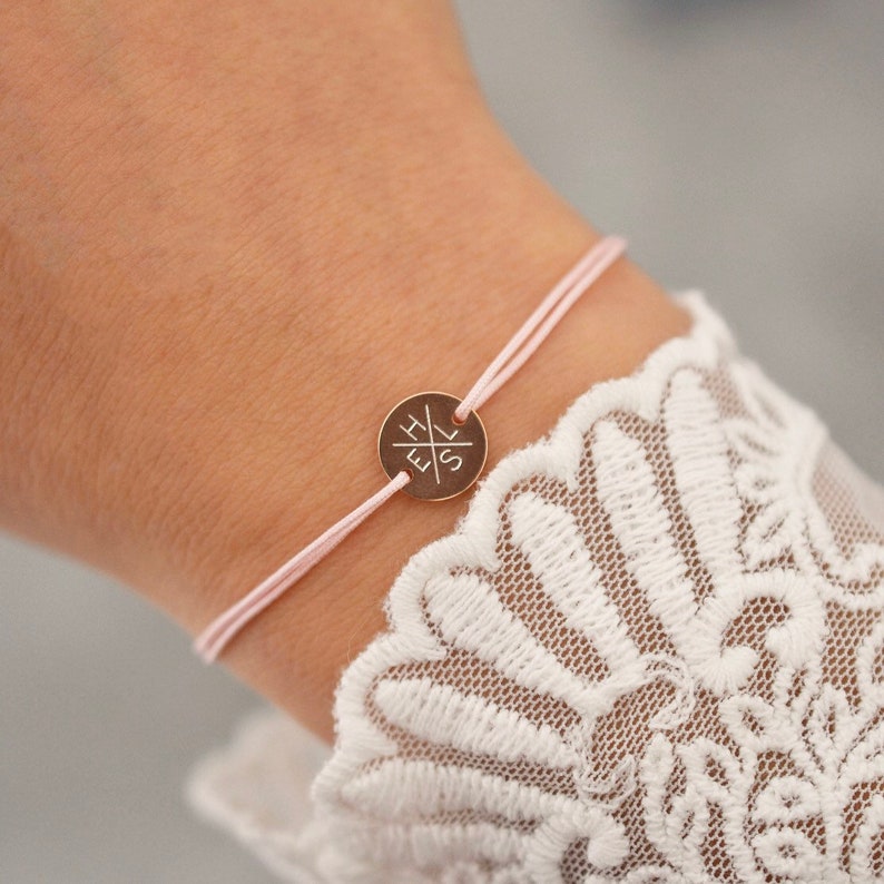 May include: A delicate pink string bracelet with a small, round, gold-colored charm. The charm has etched initials and is centered on the bracelet. The bracelet is worn on a wrist with a white lace sleeve.