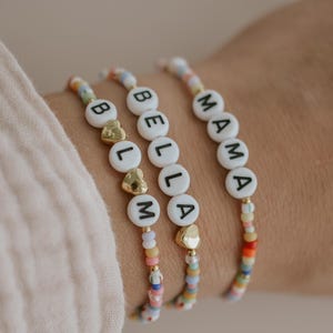 May include: Three beaded bracelets spelling out "BELLA" and "MAMA" with white letter beads and gold heart accents. The bracelets have colorful beads in pastel shades.