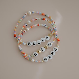 Puede incluir: Tres pulseras de cuentas con cuentas coloridas, perlas y detalles dorados. Una pulsera deletrea "BELLA", otra "MAMA", y la tercera es una mezcla de colores. Las pulseras están sobre un fondo neutro.