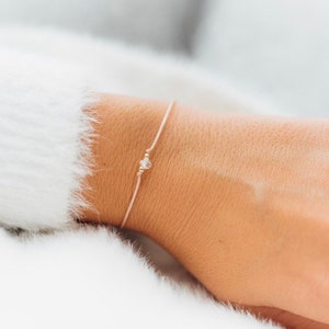 May include: Delicate light pink bracelet with a small clear crystal and gold bead accents. The bracelet is strung on a thin, light pink cord. The jewelry is simple and elegant.