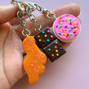 May include: A keychain set featuring three miniature food charms: a textured orange chicken nugget, a dark brown rectangular brownie topped with colorful round sprinkles, and a round cookie with pink frosting and multi-colored sprinkles.