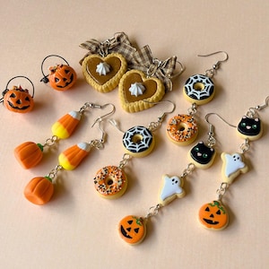Könnte beinhalten: Eine Sammlung von Halloween-Ohrhängern mit Miniatur-Anhängern, darunter orangefarbene Kürbisse, Zuckermais, herzförmige Kekse mit braunen Schleifen, Donuts mit Spinnennetz-Glasur oder Streuseln, schwarze Katzen und weiße Geister an Metallhaken.