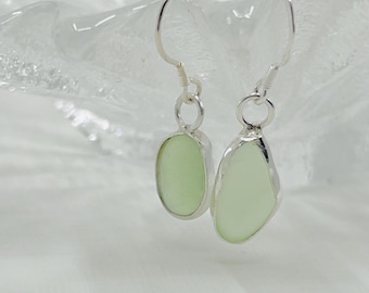 May include: A pair of earrings featuring pale green, oval-shaped sea glass set in silver. The earrings have a simple, elegant design with a hook closure. The sea glass has a smooth, frosted appearance, and the silver setting adds a touch of shine.