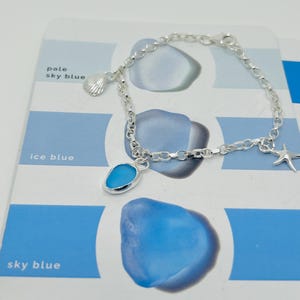 Könnte beinhalten: Ein silbernes Armband mit einer Muschel, einem blauen Seeglas-Anhänger und einem Seestern-Anhänger. Das Armband liegt auf einer blau-weißen Farbmusterkarte mit dem Text "pale sky blue", "ice blue" und "sky blue".
