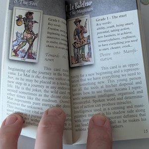 May include: An open guidebook featuring tarot card illustrations and descriptive text. The left page displays Le Mat, The Fool, and the right page displays Le Bateleur, The Magician. The pages contain printed English text detailing the meanings of these cards.