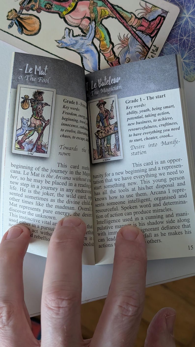 May include: An open guidebook featuring tarot card illustrations and descriptive text. The left page displays Le Mat, The Fool, and the right page displays Le Bateleur, The Magician. The pages contain printed English text detailing the meanings of these cards.
