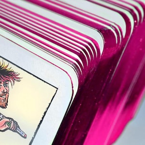 May include: A deck of tarot cards with metallic magenta edges fanned out to reveal a card featuring a colorful illustration of a person with a spiky hairstyle, a beard, and tattoos, carrying a long staff over their shoulder against a light background.