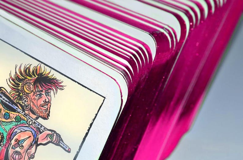 May include: A deck of tarot cards with metallic magenta edges fanned out to reveal a card featuring a colorful illustration of a person with a spiky hairstyle, a beard, and tattoos, carrying a long staff over their shoulder against a light background.