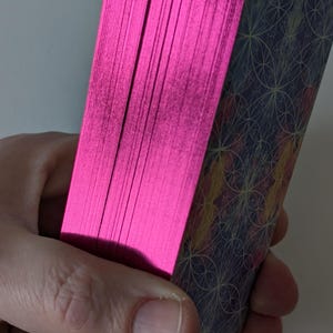 May include: A deck of rectangular cards with metallic hot pink edges. The back of the cards features a dark blue background with a repeating geometric pattern of thin, light-colored intersecting circles and lines in yellow and orange hues.