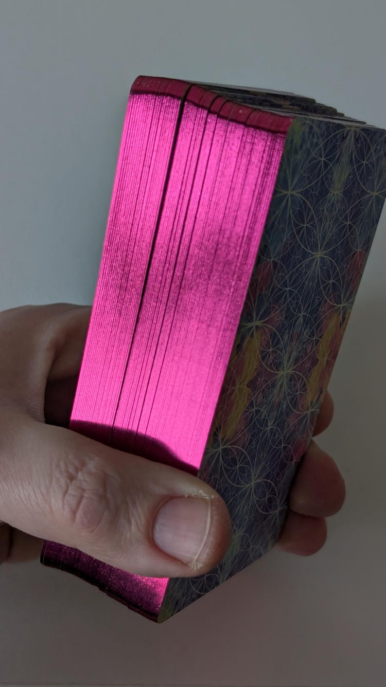 May include: A deck of rectangular cards with metallic hot pink edges. The back of the cards features a dark blue background with a repeating geometric pattern of thin, light-colored intersecting circles and lines in yellow and orange hues.