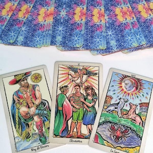 May include: A deck of tarot cards with blue, pink, and yellow geometric patterns on the back. Three cards are face up: Roy de Deniers showing a seated figure, L'Amoureux depicting three people under an angel, and La Lune featuring a moon and animals.