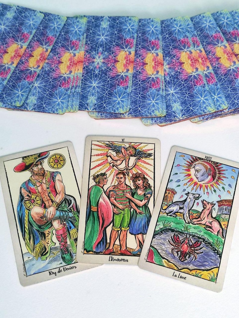 May include: A deck of tarot cards with blue, pink, and yellow geometric patterns on the back. Three cards are face up: Roy de Deniers showing a seated figure, L'Amoureux depicting three people under an angel, and La Lune featuring a moon and animals.