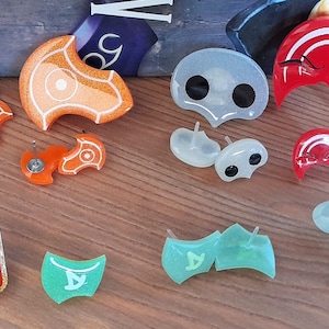 Könnte beinhalten: Sortiment an bunten Harzohrringen und -nadeln mit geometrischen Formen, einem Totenkopf und einem Halbmond. Die Ohrringe sind in Orange, Weiß, Rot und Grün. Die Nadeln sind in Rot und Grün.