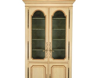 May include: A tall, cream-colored wooden cabinet with two glass doors featuring a wire mesh design. The cabinet has two lower doors with decorative carvings and brass hardware.